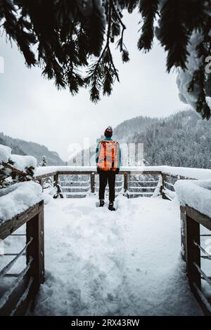 Man Tourist und Kletterer mit orangem Rucksack, der am Aussichtspunkt (Ort) steht, mit fantastischem Blick auf verschneite Berge im kalten Winter. Backpacker Stockfoto