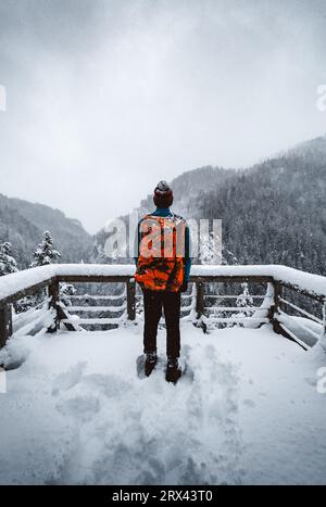 Man Tourist und Kletterer mit orangem Rucksack, der am Aussichtspunkt (Ort) steht, mit fantastischem Blick auf verschneite Berge im kalten Winter. Backpacker Stockfoto
