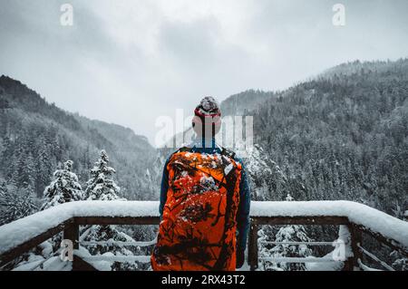 Man Tourist und Kletterer mit orangem Rucksack, der am Aussichtspunkt (Ort) steht, mit fantastischem Blick auf verschneite Berge im kalten Winter. Backpacker Stockfoto