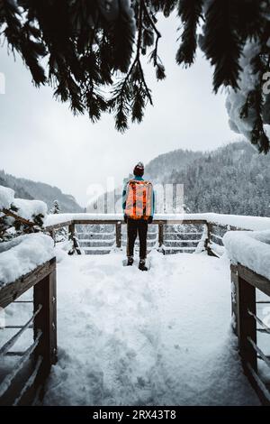 Man Tourist und Kletterer mit orangem Rucksack, der am Aussichtspunkt (Ort) steht, mit fantastischem Blick auf verschneite Berge im kalten Winter. Backpacker Stockfoto