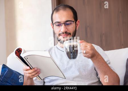 Bärtiger arabischer Mann trinkt Kaffee, während er Buch liest und auf der Couch im Wohnzimmer sitzt Stockfoto