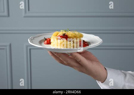 Der Kellner hielt einen Teller mit leckerem Capellini mit Tomaten und Käse in der Nähe der grauen Wand, Schließung. Exquisite Präsentation eines Nudelgerichts Stockfoto