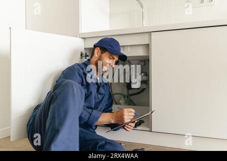 Professioneller Klempner untersucht nach der Installation ein Siphonrohr am Spülbecken und macht sich Notizen Stockfoto