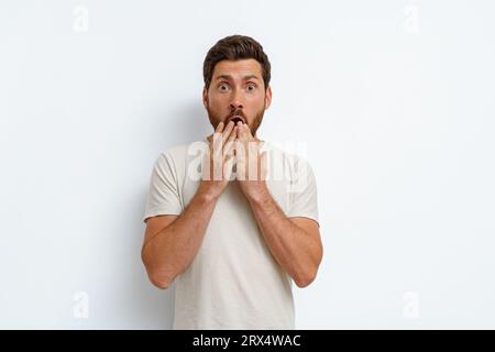 Der gutaussehende Mann im T-Shirt bedeckt seinen Mund mit den Händen mit einem schockierten Ausdruck auf weißem Hintergrund Stockfoto