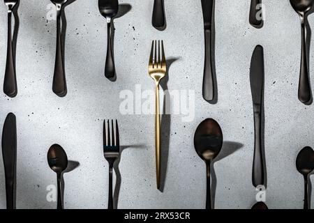 Eine goldene Gabel unter schwarzem Besteck. Neutraler grauer Betonhintergrund Stockfoto
