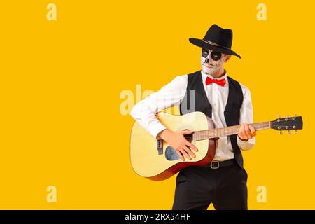 Junger Mann mit gemaltem Schädel, der Gitarre auf gelbem Hintergrund spielt. Feierlichkeiten zum mexikanischen Todestag (El Dia de Muertos) Stockfoto