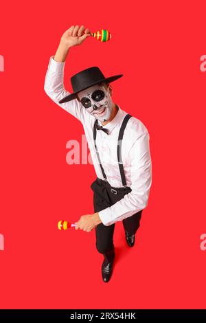 Junger Mann mit gemaltem Schädel und Maracas auf rotem Hintergrund. Feierlichkeiten zum mexikanischen Todestag (El Dia de Muertos) Stockfoto