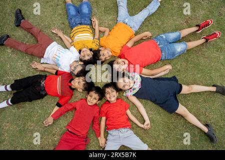 Gruppe glücklicher indischer Kinder, die bunte Tücher tragen, die im Sommerpark auf Gras liegen, verspielte asiatische Kinder, die im Garten im Freien zusammen spielen. Anzeigen Stockfoto