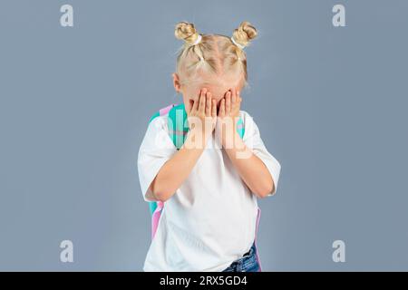 Zurück zur Schule. Kleines Schulmädchen mit Rucksack, der ihr Gesicht mit den Händen bedeckt Stockfoto