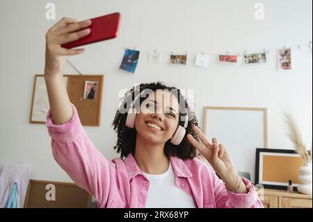 Glückliches Mädchen im Teenageralter, das Friedenszeichen zeigt und Selfie über das Smartphone nimmt Stockfoto