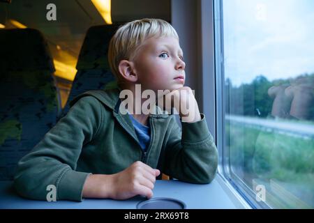 Nachdenklicher Junge mit der Hand auf dem Kinn, der im Zug durch das Fenster schaut Stockfoto