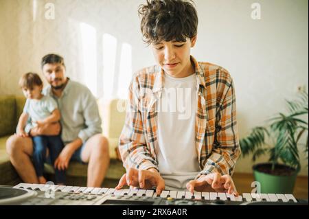 Junge, der Klavier spielt, während Vater und Bruder im Hintergrund sitzen Stockfoto