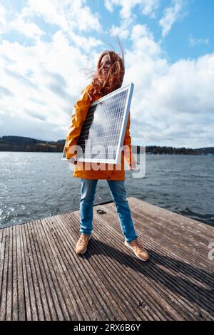 Frau mit Sonnenkollektor steht auf dem Pier unter bewölktem Himmel am See Stockfoto