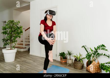 Frau, die zu Hause eine VR-Brille benutzt und das Knie dehnt Stockfoto