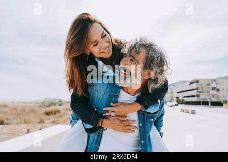 Glücklicher älterer Mann, der eine Frau mit Huckepack zum Strand trägt Stockfoto