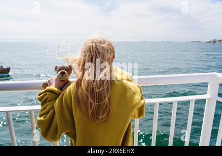 Blondes Mädchen mit Teddybär in der Nähe eines Geländes in der Fähre, die auf See fährt Stockfoto