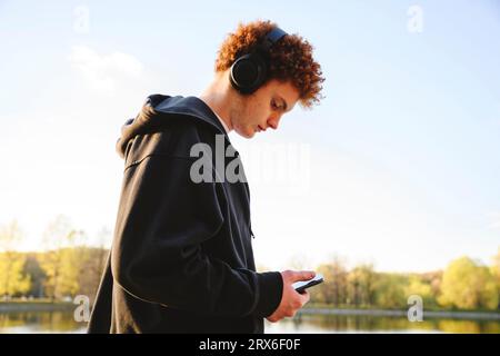 Teenager-Junge, der das Mobiltelefon nutzt und Musik über Kopfhörer im Park hört Stockfoto