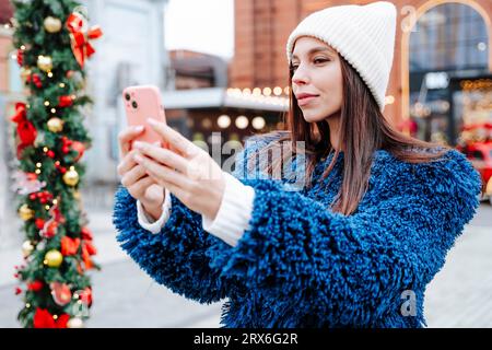 Frau, die Strickmütze trägt, nimmt Selfie auf dem Weihnachtsmarkt Stockfoto