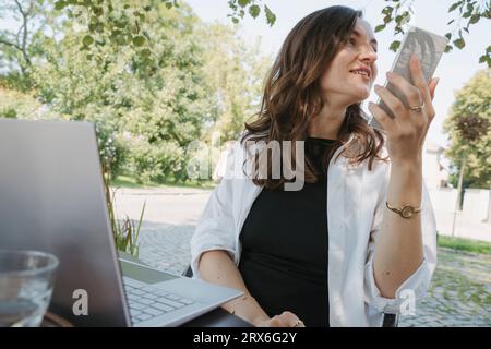 Lächelnde Geschäftsfrau, die Voicemail über das Smartphone am Tisch im Garten aufnimmt Stockfoto