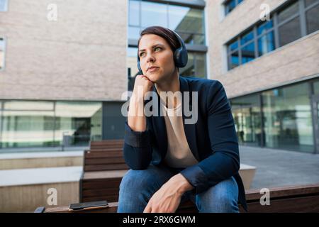 Geschäftsfrau mit der Hand am Kinn, die Musik über kabellose Kopfhörer hört Stockfoto