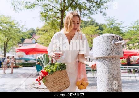 Frau mit Tüten voller Lebensmittel, die Wasser aus dem Trinkbrunnen holen Stockfoto