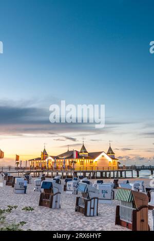 Deutschland, Mecklenburg-Vorpommern, Ahlbeck, Strandliegen mit Kapuze bei Sonnenuntergang mit Pier und Badehaus im Hintergrund Stockfoto