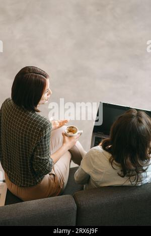 Zwei Mitarbeiterinnen, die während einer Pause über problematische Aufgaben bei der Arbeit diskutieren. Kurzhaariges Mädchen, das beim Reden eine Tasse Kaffee hält Stockfoto