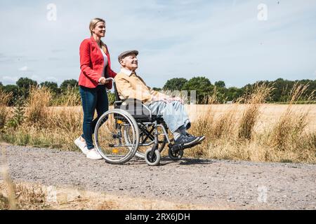 Frau, die einen älteren Mann schiebt, der im Rollstuhl sitzt, auf dem Feld Stockfoto