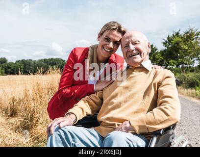 Glückliche Frau mit einem älteren Mann, der an einem sonnigen Tag im Rollstuhl sitzt Stockfoto