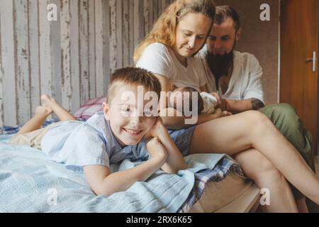 Lächelnder Sohn, der neben den Eltern liegt und eine neugeborene Tochter zu Hause im Bett hat Stockfoto