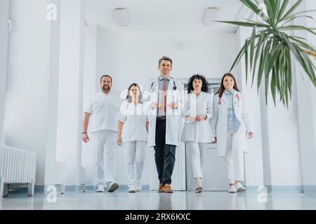 Eine selbstbewusste Gruppe von Ärzten, einschließlich einer Fachärztin, geht durch einen Krankenhausgang. Sie tragen medizinische Uniformen und Labormäntel und das Lächeln Stockfoto