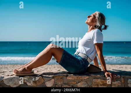 Frau mit geschlossenen Augen sitzt auf der Promenade am Strand Stockfoto