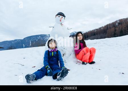 Zwei Kinder sitzen neben dem Schneemann, den sie auf dem schneebedeckten Berg gemacht haben Stockfoto