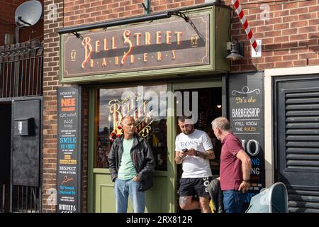 Drei Männer stehen vor dem Bell Street Barbers Shop mit seinen traditionellen Schildern und rot-weißen Stangen in North Shields, North Tyneside, UK. Stockfoto