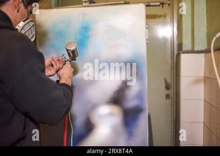 Ein hispanischer junger Mann, der eine Schutzmaske trägt, testet in seiner Werkstatt eine Spritzpistole Stockfoto