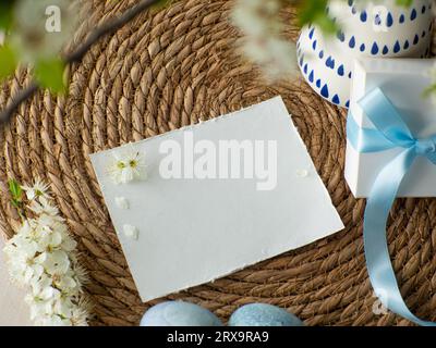 Blumenstrauß mit Kirschblüten in Vase, Geschenkbox mit blauem Bogen und Papierkarte mit Kopierraum auf Strohhintergrund. Frauen, Männer und Muttertagsgeschenk. Konzept Stockfoto