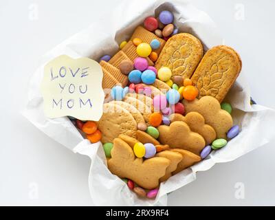 Verschiedene Kekse und Süßigkeiten im Korb mit "Love You mom" -Zettel. Muttertagskonzept. Handgemachtes Pflegespaket, saisonale Geschenkbox mit Süßigkeiten, Dragee Stockfoto