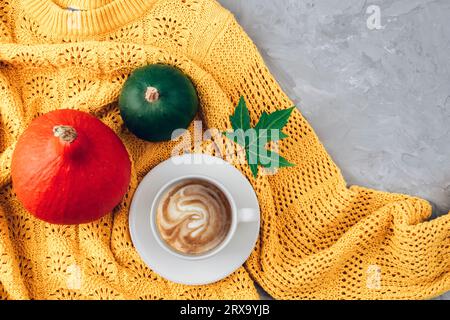 Herbstflachlage mit orangefarbenen und grünen Kürbissen und Kaffeetasse auf gelbem Pullover auf grauem Betonhintergrund. Draufsicht, Kopierbereich. Stockfoto