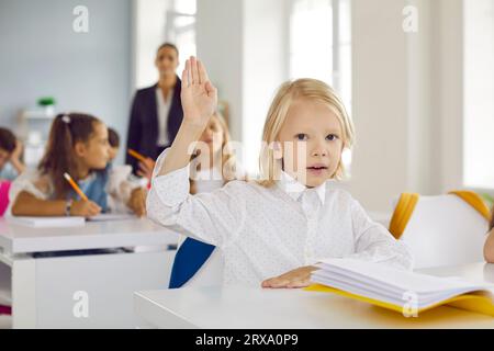 Der kleine blonde Grundschuljunge in einem weißen Hemd, der die Hand hebt, sitzt im Klassenzimmer am Schreibtisch Stockfoto