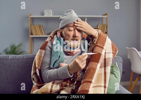 Kranker älterer Mann, der seine Temperatur in einer Decke mit Kopfschmerzen überprüft. Stockfoto