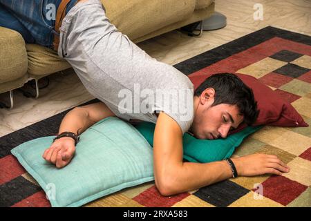 Foto eines jungen und gutaussehenden Mannes, der in einer seltsamen Position auf dem Boden mit einem Kissen unter dem Kopf ruht, vielleicht betrunken oder sehr müde Stockfoto