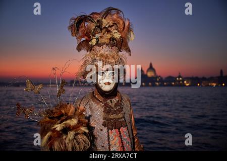 Genießen Sie venezianische Traditionen, während die Sonne über San Giorgio Maggiore untergeht, wo Masken und Magie sich im zauberhaften Karneval von 2023 vereinen. Stockfoto