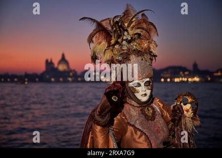 Genießen Sie venezianische Traditionen, während die Sonne über San Giorgio Maggiore untergeht, wo Masken und Magie sich im zauberhaften Karneval von 2023 vereinen. Stockfoto