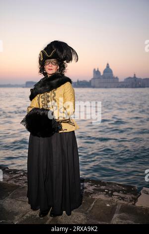 Genießen Sie venezianische Traditionen, während die Sonne über San Giorgio Maggiore untergeht, wo Masken und Magie sich im zauberhaften Karneval von 2023 vereinen. Stockfoto