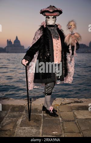 Genießen Sie venezianische Traditionen, während die Sonne über San Giorgio Maggiore untergeht, wo Masken und Magie sich im zauberhaften Karneval von 2023 vereinen. Stockfoto