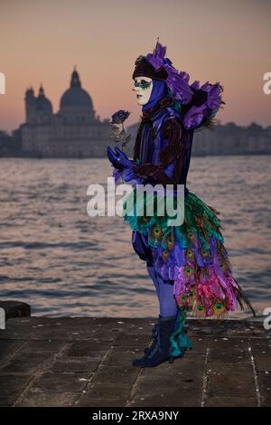 Genießen Sie venezianische Traditionen, während die Sonne über San Giorgio Maggiore untergeht, wo Masken und Magie sich im zauberhaften Karneval von 2023 vereinen. Stockfoto