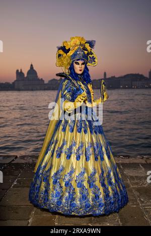 Genießen Sie venezianische Traditionen, während die Sonne über San Giorgio Maggiore untergeht, wo Masken und Magie sich im zauberhaften Karneval von 2023 vereinen. Stockfoto