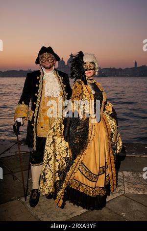 Genießen Sie venezianische Traditionen, während die Sonne über San Giorgio Maggiore untergeht, wo Masken und Magie sich im zauberhaften Karneval von 2023 vereinen. Stockfoto