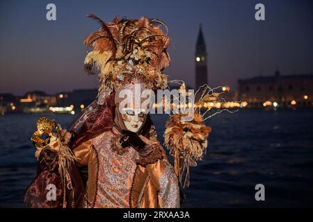 Genießen Sie venezianische Traditionen, während die Sonne über San Giorgio Maggiore untergeht, wo Masken und Magie sich im zauberhaften Karneval von 2023 vereinen. Stockfoto