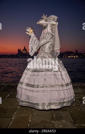 Genießen Sie venezianische Traditionen, während die Sonne über San Giorgio Maggiore untergeht, wo Masken und Magie sich im zauberhaften Karneval von 2023 vereinen. Stockfoto